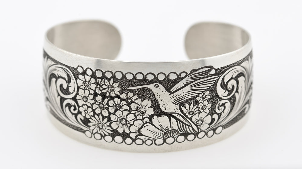 Silver cuff bracelet with engraved design of a hummingbird and flowers on a white background