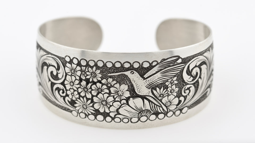 Silver cuff bracelet with engraved design of a hummingbird and flowers on a white background