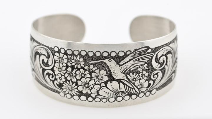 Silver cuff bracelet with engraved design of a hummingbird and flowers on a white background