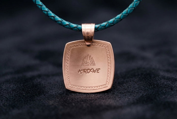 Bronze pendant with engraved text on a turquoise leather cord against a dark background
